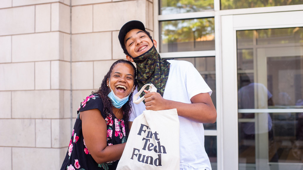 Two people smiling and embracing outside a building, one holding a tote bag with text "fix tech fund." both are wearing face masks.