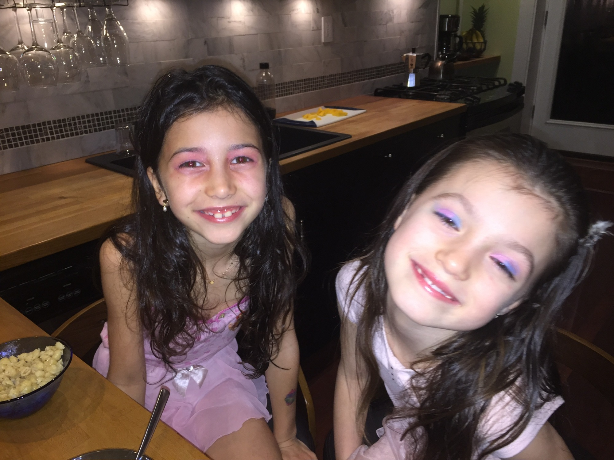 Two young girls with colorful makeup smiling at a kitchen counter with a bowl of popcorn nearby.