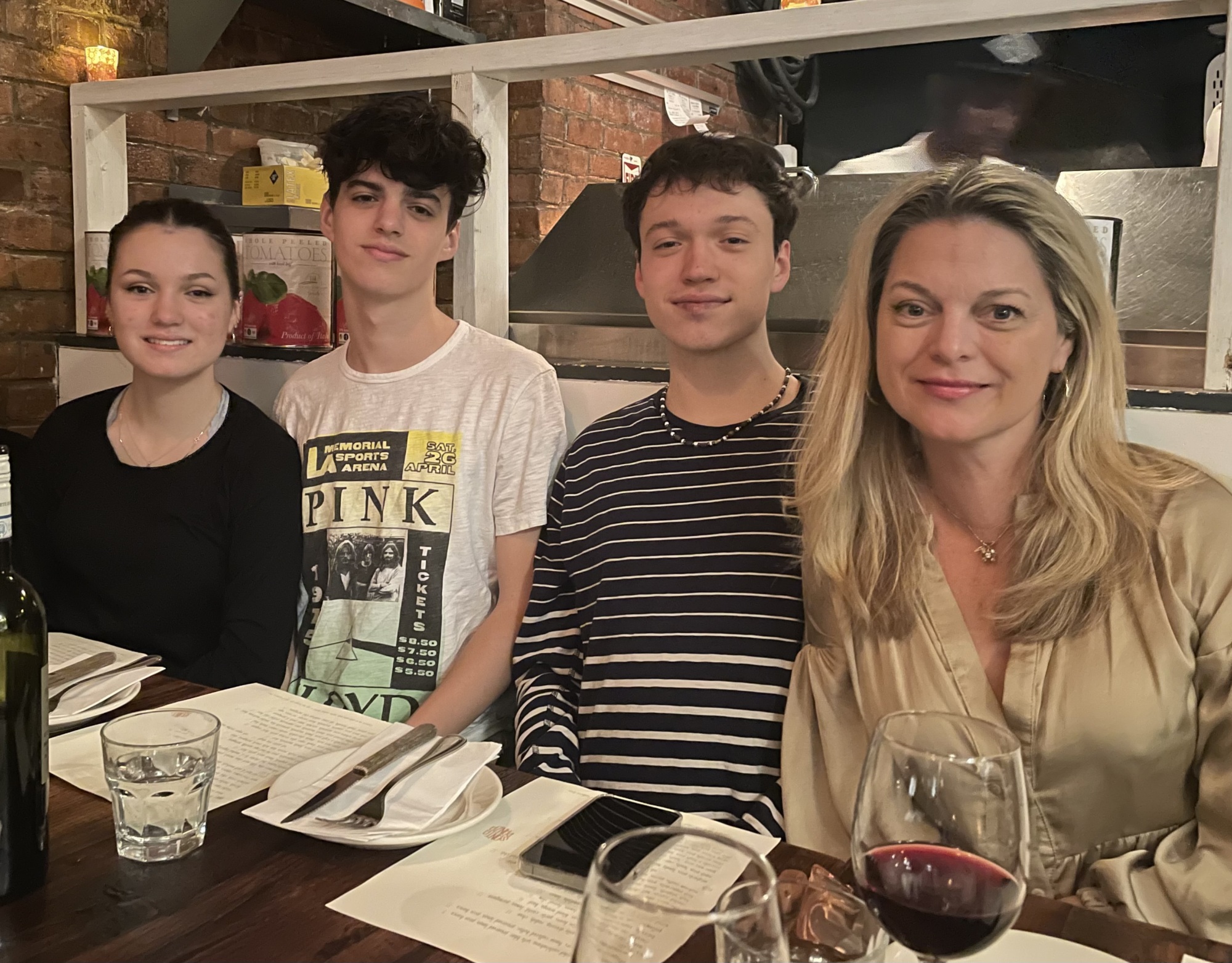 A mother and three teenagers sitting at a restaurant table, smiling at the camera with glasses and a wine bottle on the table.