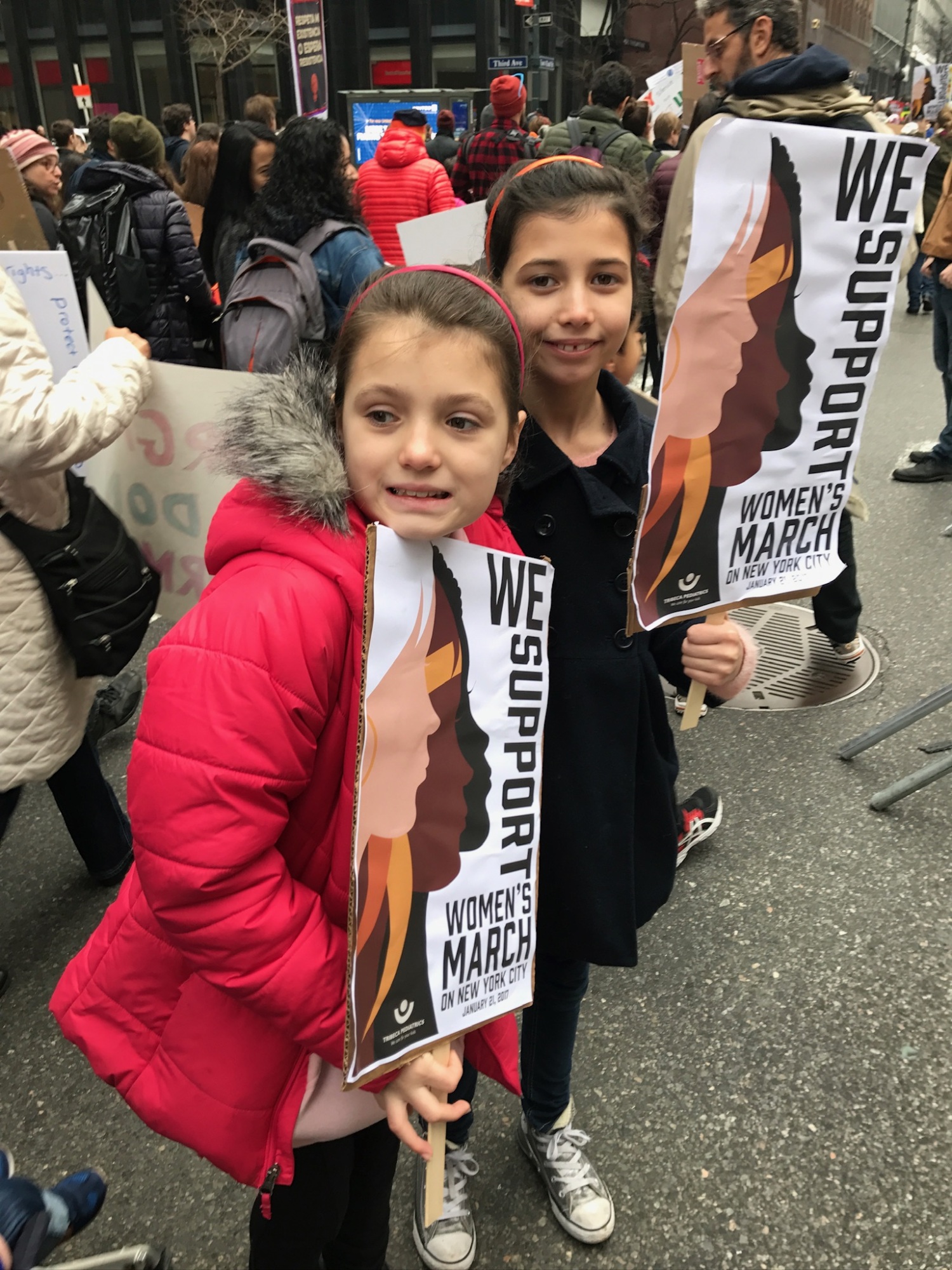 Two young girls holding "we support women's march" banners at a crowded street demonstration.
