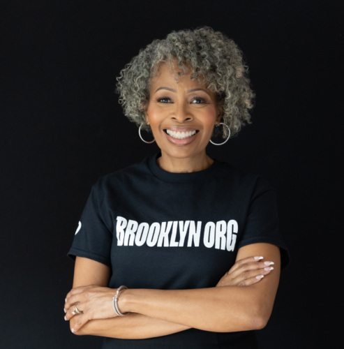 Portrait of a smiling Black woman with curly gray hair, wearing a "Brooklyn Org" t-shirt, standing with arms crossed on a black background.