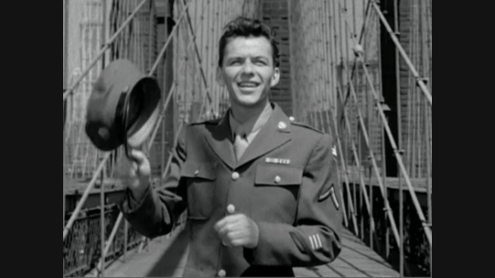 A smiling man in a military uniform holds his hat in one hand, standing in front of a large suspension bridge under construction.