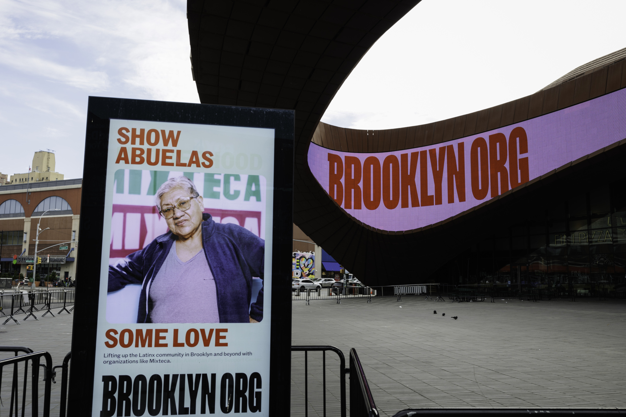 A poster showing an older woman saying "Show Abuelas Some Love" in front of the Barclays Center with a large "BROOKLYN.ORG" sign displayed on the building in Brooklyn, New York.
