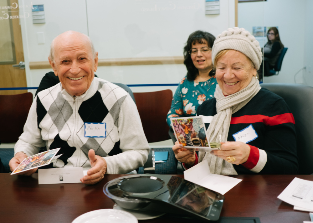 Two senior adults, a joyful bald man and a smiling woman in a gray beanie, sit at a table holding brochures, surrounded by people in a seminar room.