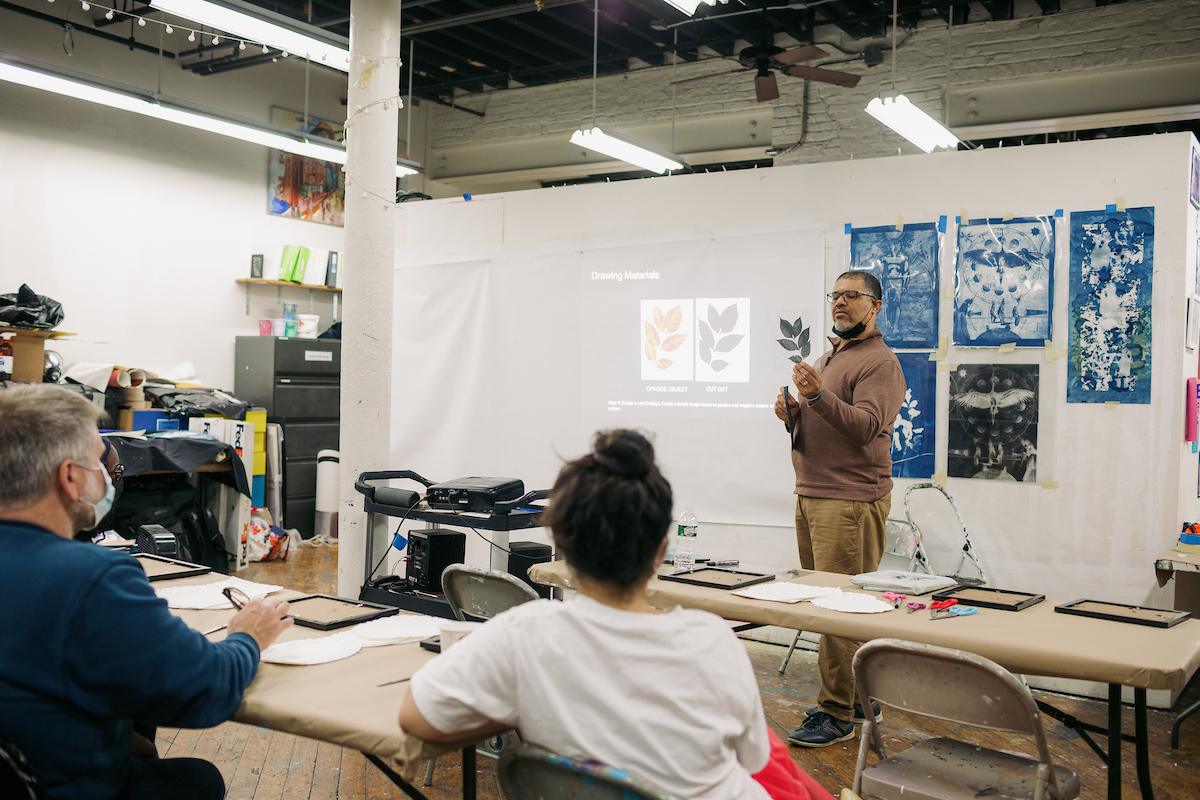 An instructor stands near a presentation screen showing leaf illustrations, teaching a small group of people seated at desks in an art classroom.