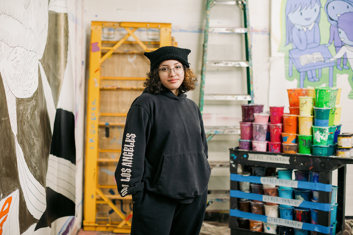 A person wearing a black beanie and hoodie stands in an art studio, surrounded by colorful paint cans stacked on a wooden frame and artworks on the walls.