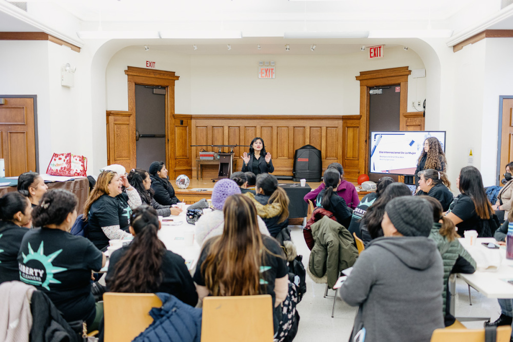 A group of people is seated in a room with wooden doors, listening to a person speaking at the front. A presentation screen is visible, and the attendees appear engaged.