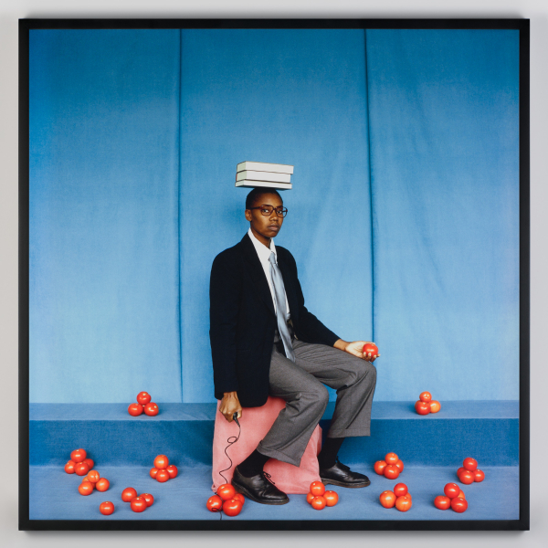A person in formal attire balances three books on their head while seated on a pink stool, holding a corded phone receiver and surrounded by scattered red tomatoes against a blue background.