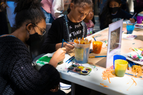 Children engaged in painting activities at a table, with art supplies and cups of paint scattered around. One child is wearing a black mask and the other child is focused on their painting.