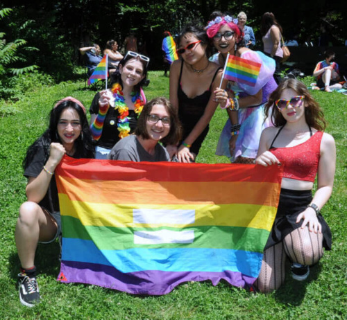 A group of people wearing colorful outfits, gathered outdoors holding LGBTQ+ flags and a rainbow flag with an equals sign, smiling at the camera.