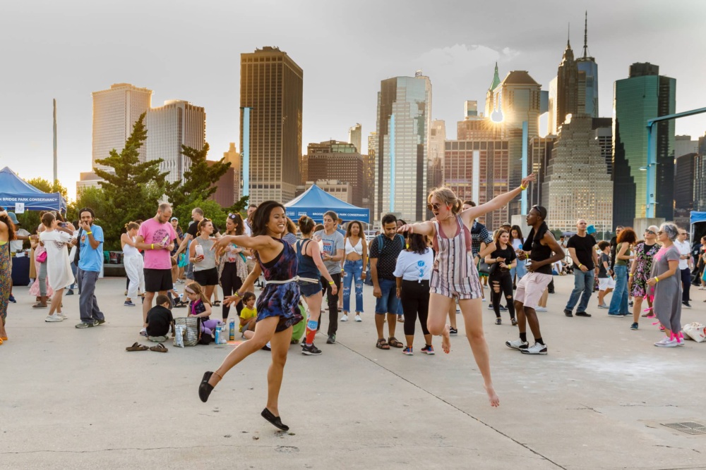 A group of people is gathered outdoors in a city plaza with skyscrapers in the background. Two women in the foreground are dancing energetically.
