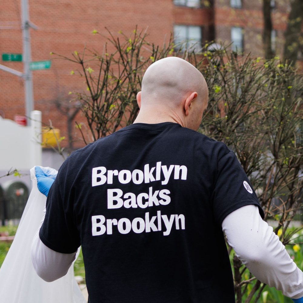 Person with a bald head wearing a "Brooklyn Backs Brooklyn" shirt, holding a trash bag and standing in front of some bushes and buildings on a city street.