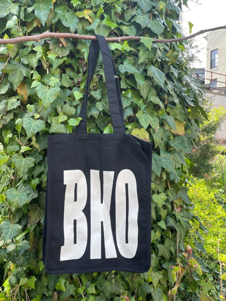 A black tote bag with large white letters "BKO" hangs from a branch, with a background of green ivy.