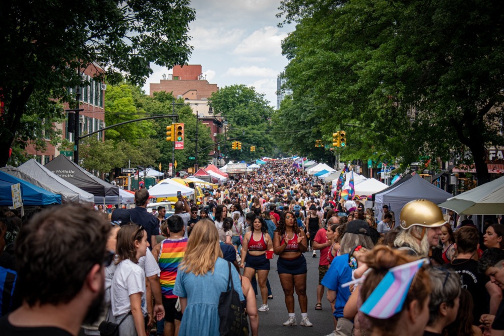A large crowd participating in an outdoor street festival next to vendor booths, with people wearing colorful clothing and rainbow flags visible. Trees and buildings line the street.