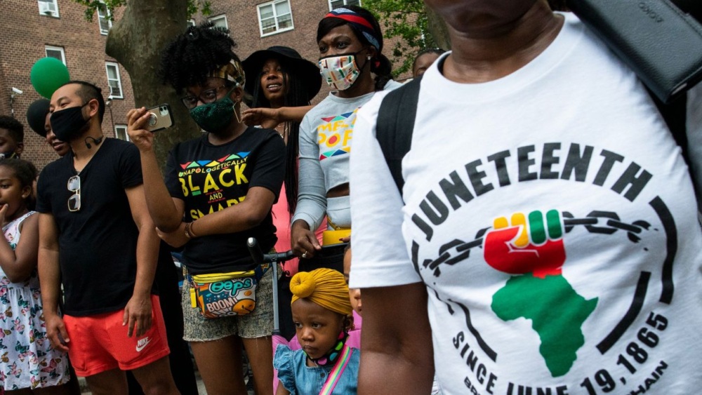 A diverse group of people, including children, gather outdoors. A woman in the foreground wears a shirt that reads "Juneteenth Since June 19, 1865" with a design of a fist and chain.