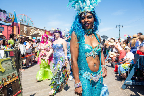 A group of people dressed as mermaids in colorful costumes walk in a parade along a boardwalk filled with spectators.