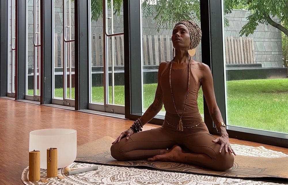 A person wearing brown yoga attire sits cross-legged on a mat inside a room with large windows, eyes closed, and hands resting on knees. Outside, trees and grass are visible.