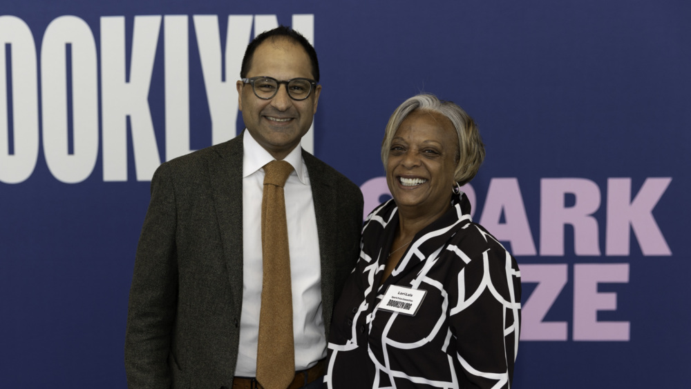 Two smiling adults, a man and a woman, stand in front of a blue backdrop with "Brooklyn Org Spark Prize" text, man in a suit and glasses, woman in a black and white patterned top.