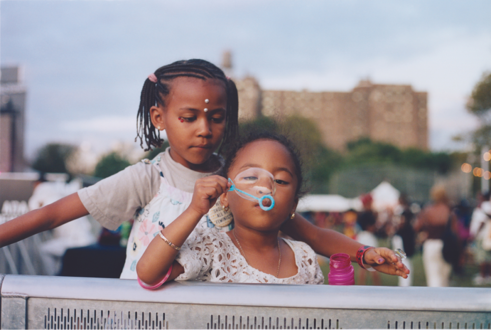 Two young children at an outdoor event; one child is blowing bubbles while the other stands close behind, looking on. Urban buildings and sky are visible in the background.