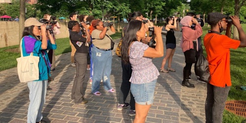 A group of people using cameras and binoculars outdoors, focusing on something in the distance.