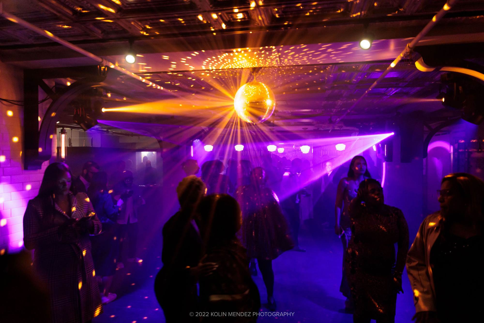 People dancing under a disco ball in a dimly lit club with colorful lights and haze.