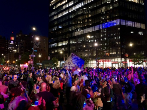 A large crowd of people wearing illuminated headphones dance outside a lit-up office building at night.