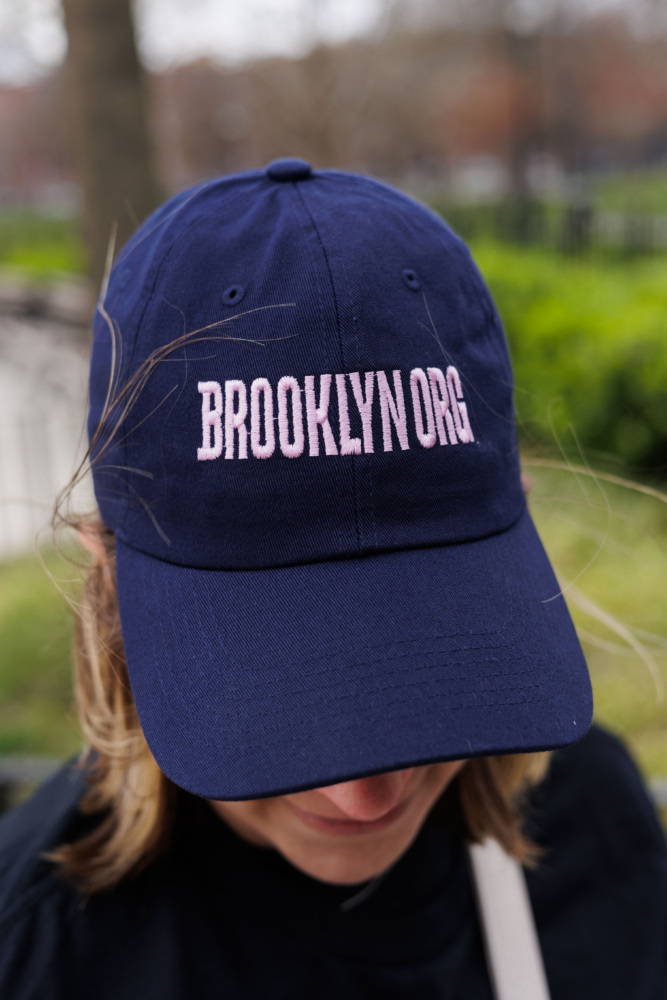 A person wearing a navy blue cap embroidered with the words "BROOKLYN.ORG" stands outdoors.