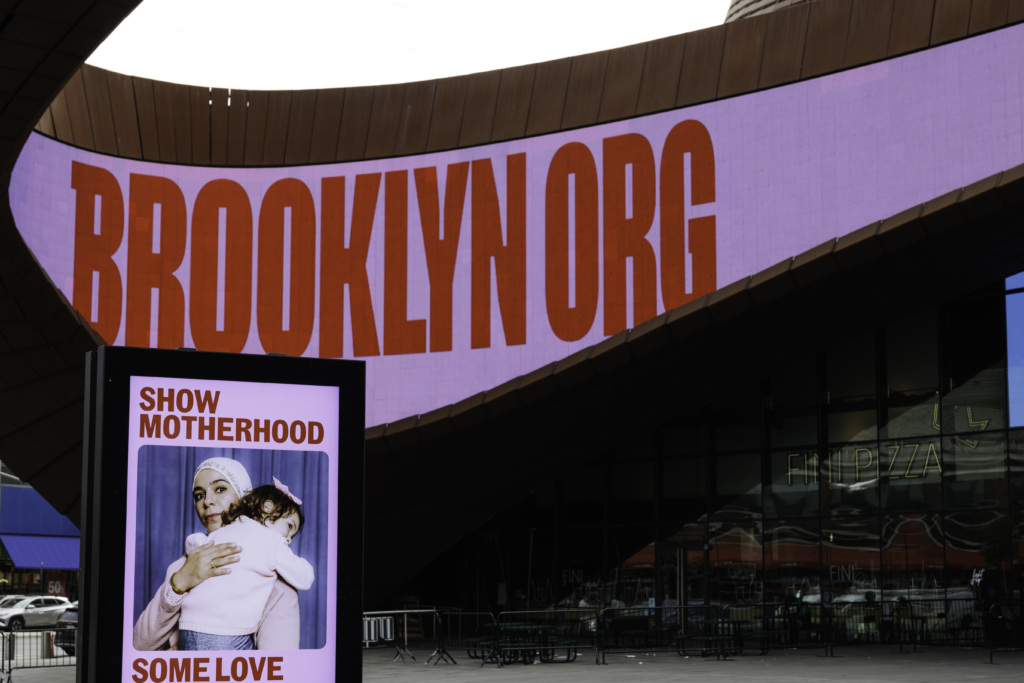 Large display reading "BROOKLYN.ORG" with a smaller sign in the foreground showing a woman holding a child and the text "SHOW MOTHERHOOD SOME LOVE" outside a building.
