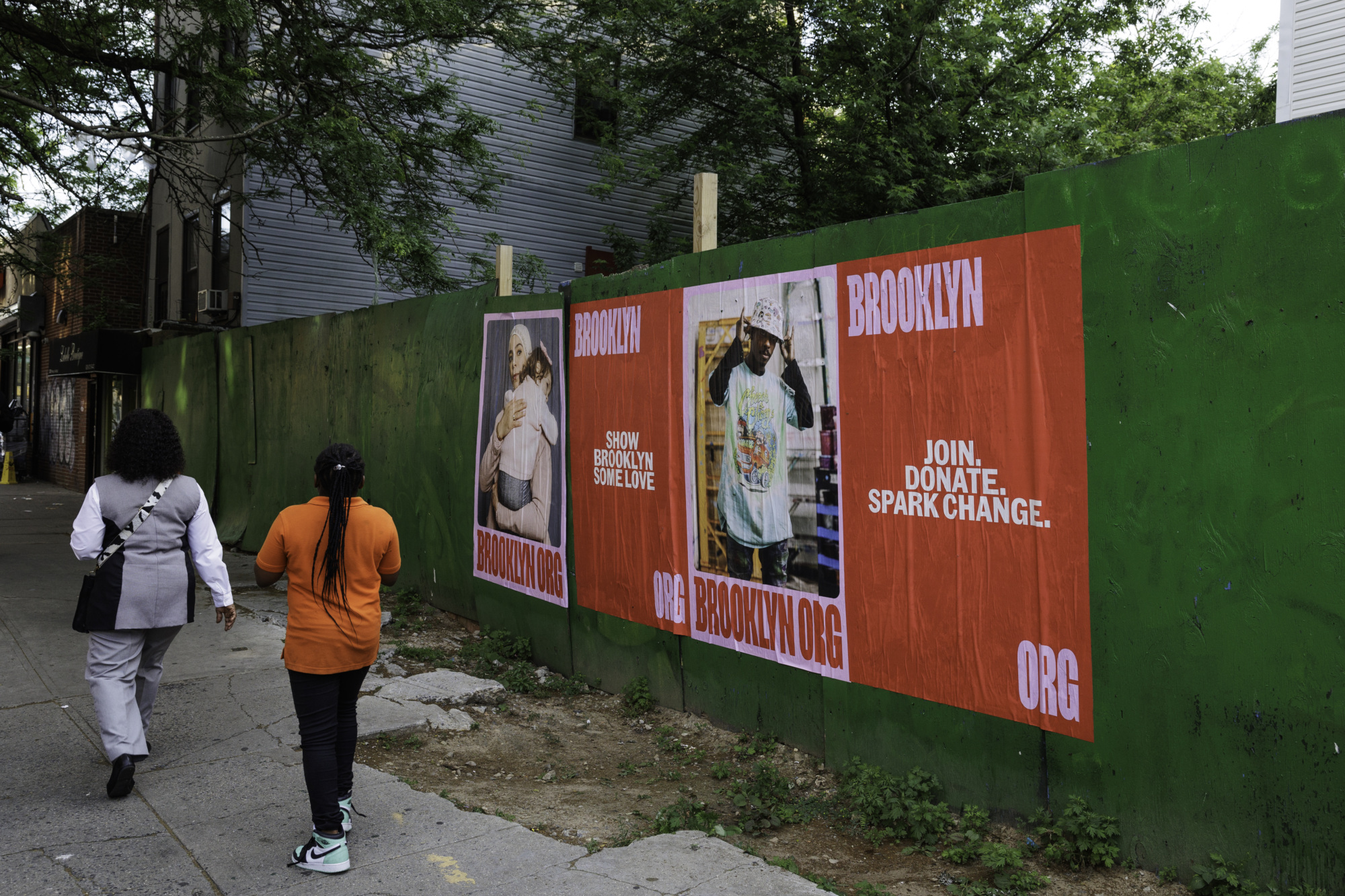 Two people walk past a green fence featuring three red posters that advertise a Brooklyn-based organization, encouraging donations and volunteer support.