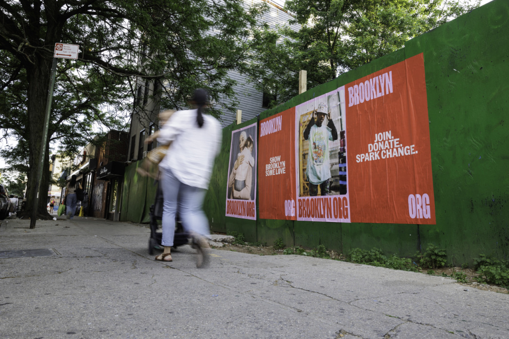 A person walks past a green construction barrier with three red posters for brooklyngo.org, promoting donations for the organization. The posters feature different images and messages.