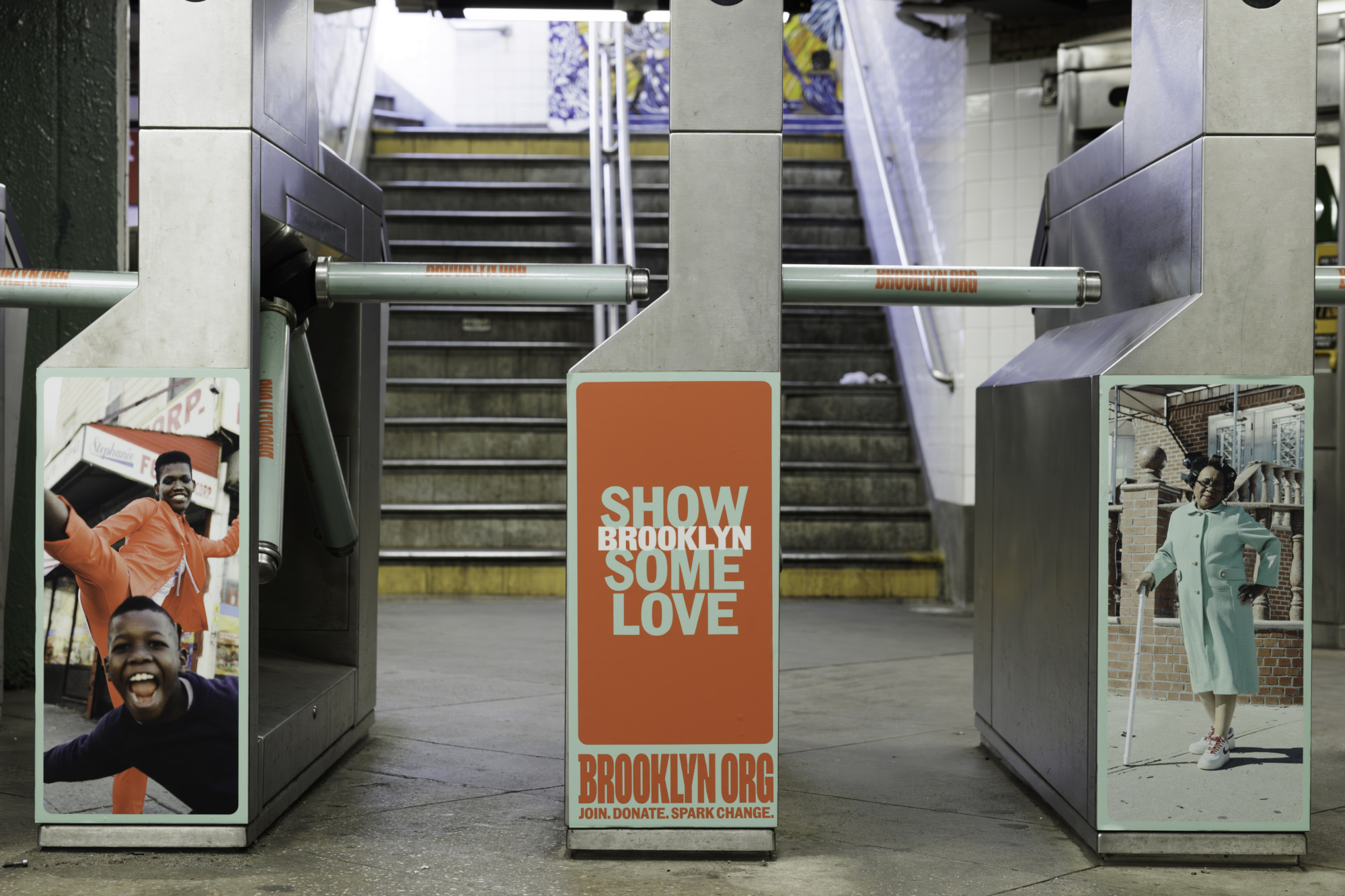 Subway turnstiles with signs that read "Give to Brooklyn." Two posters show smiling people. Stairs leading to an upper level are in the background.