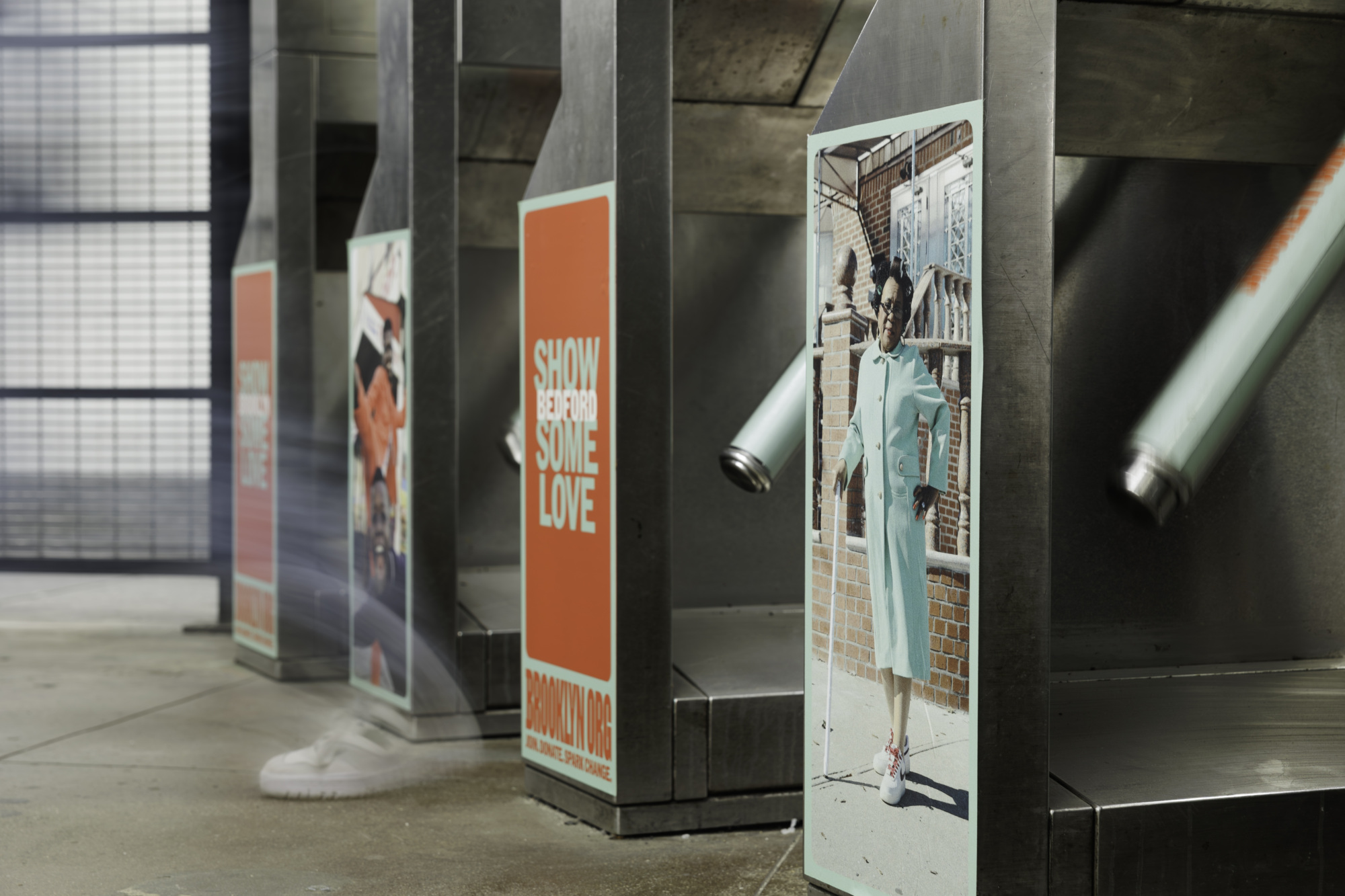 A public space with turnstiles, partially open. Each turnstile has a poster; one poster reads "SHOW BE KINDNESS SOME LOVE," and another shows a person in a light blue outfit walking.