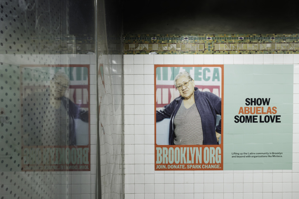 A poster on a tiled wall featuring a person with text urging support for grandmothers (abuelas) through donations and involvement with BROOKLYN.ORG. Reflections of the poster are seen on the left side.