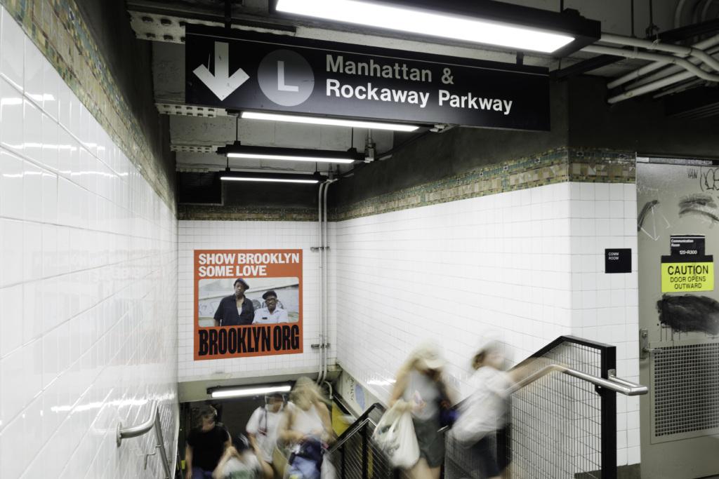 A subway station entrance with a sign for the L train to Manhattan and Rockaway Parkway. People are walking down the stairs. A poster on the wall reads "Show Brooklyn Some Love" with a website link.