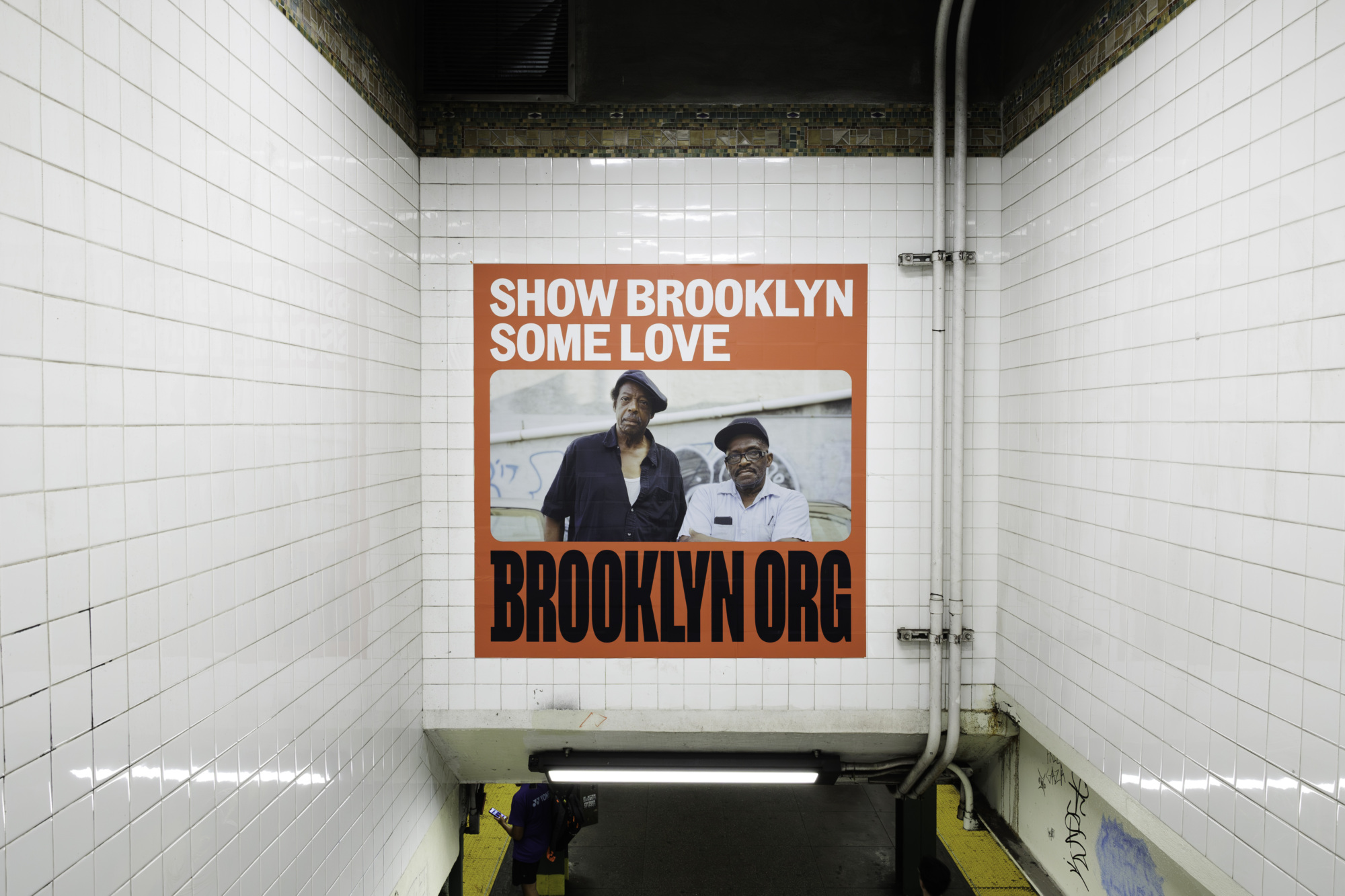 A wall poster in a subway station features two men and reads, "SHOW BROOKLYN SOME LOVE" with "GIVE TO BROOKLYN.ORG" at the bottom. The walls are tiled with a white grid pattern.