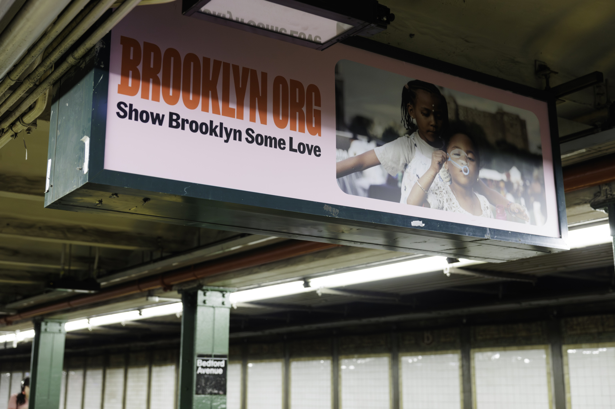 A subway platform with a sign displaying "BROOKLYN.ORG Show Brooklyn Some Love" and an image of two children, one blowing bubbles. The station name, "Bedford Avenue," is visible on the platform post, illustrating the warmth and spirit of those who give to Brooklyn.