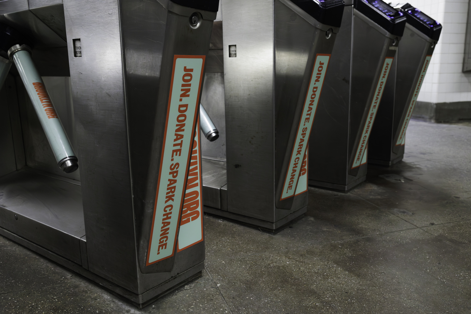 A row of subway turnstiles with orange and white advertisements reading "Join. Donate. Spark Change." and a website link on a gray tiled floor in a subway station.