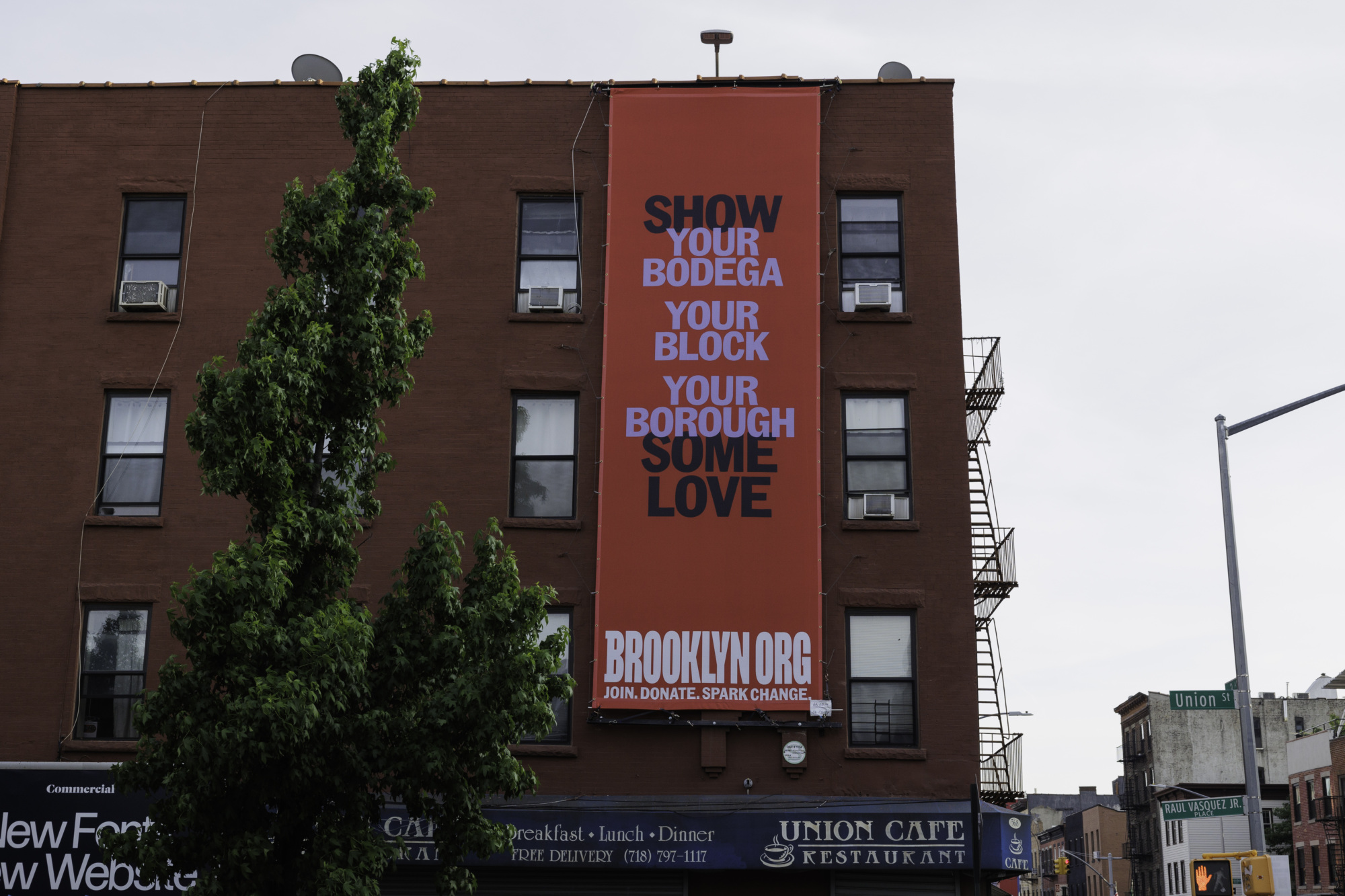 A large red banner on a building reads, "Show your bodega your block your borough some love - Give to Brooklyn at BROOKLYN.ORG Join. Donate. Spark Change." The building's street signs indicate Union Street.