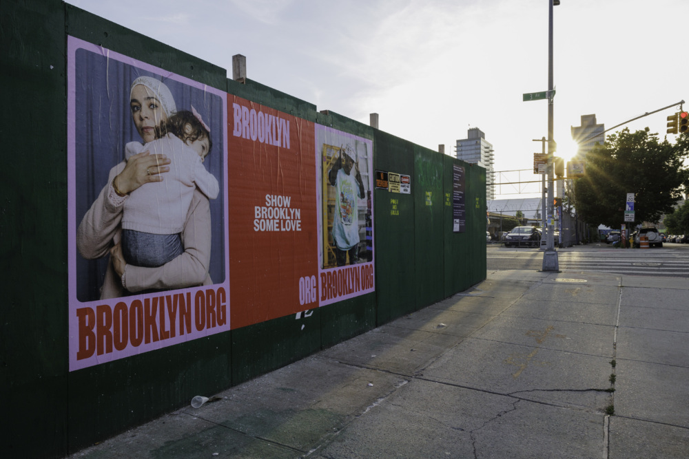 A sidewalk with posters on a green wall, one showing a person holding a child, and the other with the text "SHOW BROOKLYN SOME LOVE." The street is in an urban area with buildings and a traffic light.