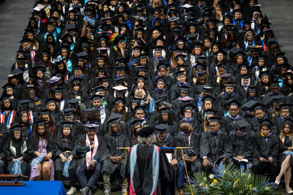 A large group of graduates in caps and gowns sit in rows while a speaker in academic regalia addresses them from the front at a graduation ceremony.
