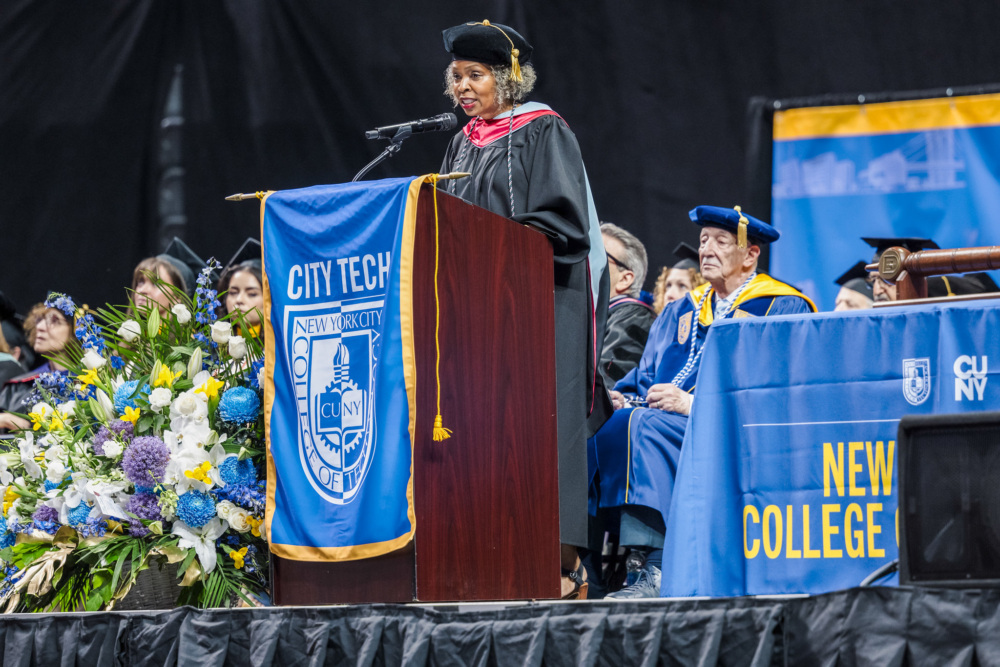 A person in academic regalia speaks at a podium during a university graduation ceremony. Flowers are on the left, and several faculty members in gowns are seated on the right.