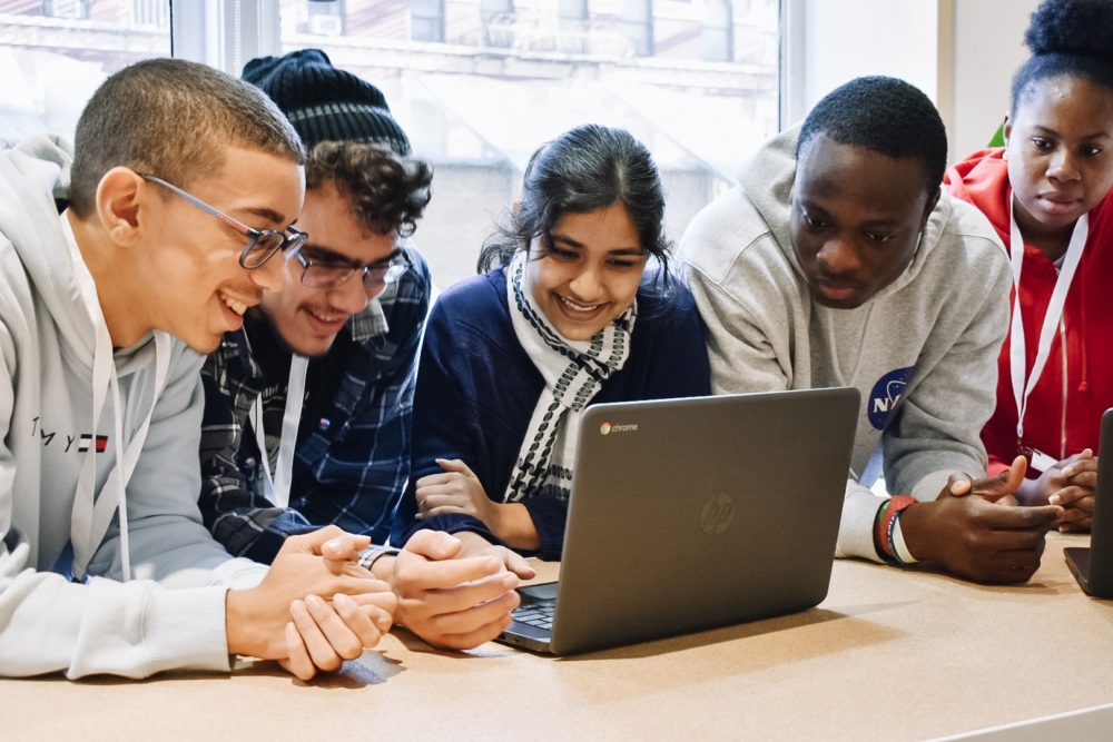 A group of five people sit closely together around a table, smiling and looking at a laptop screen.