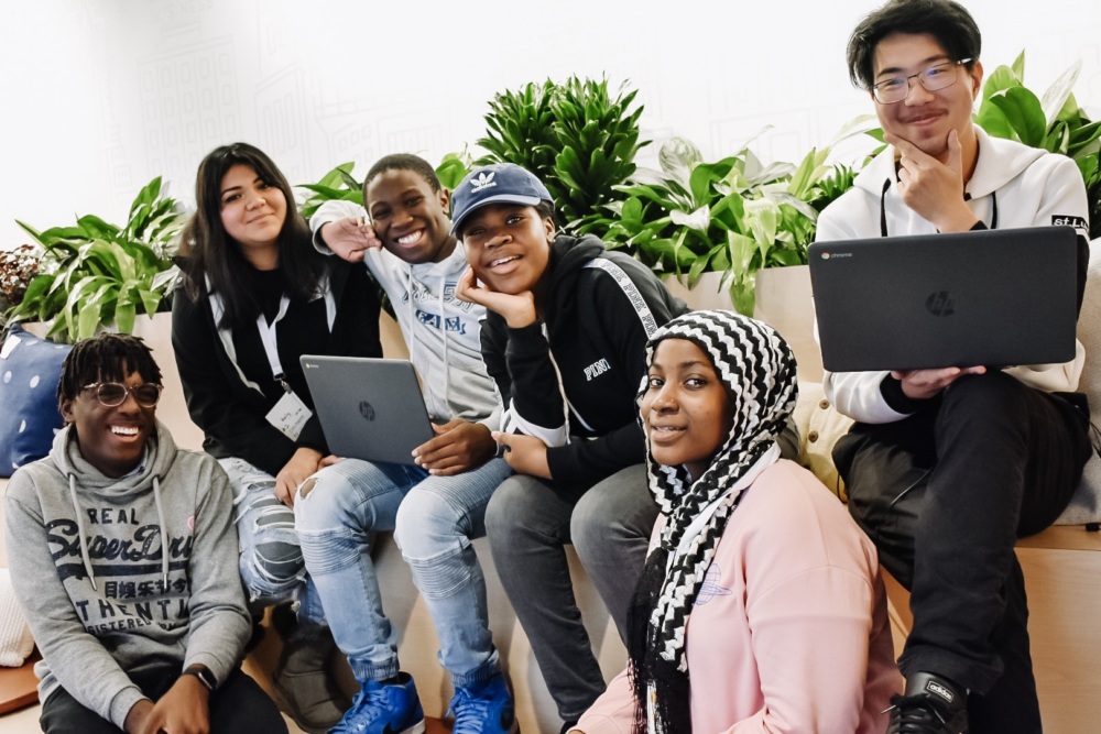 A group of six diverse individuals, some holding laptops, sit and pose together in front of indoor plants, smiling at the camera.