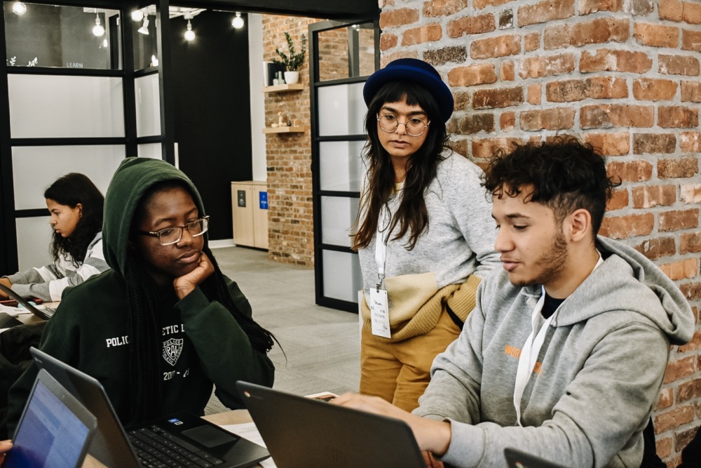 Three people work on laptops while another individual looks on, in a room with brick walls and glass partitions.