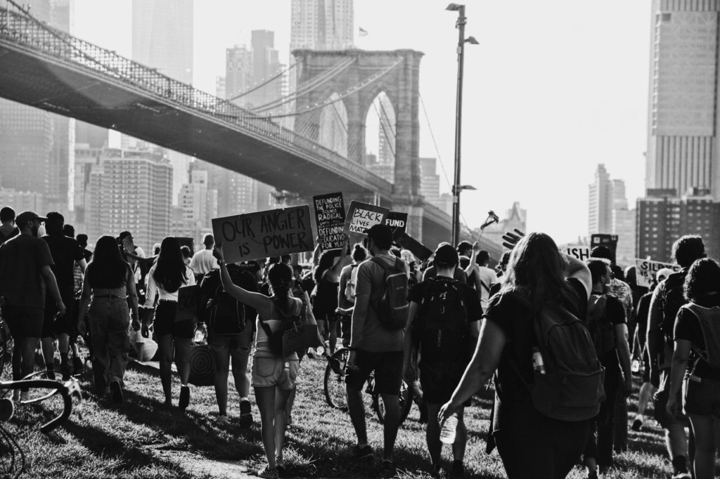 Black and white image of a group of people walking together near a bridge, holding signs with messages, one sign reads "Our Anger is Power.