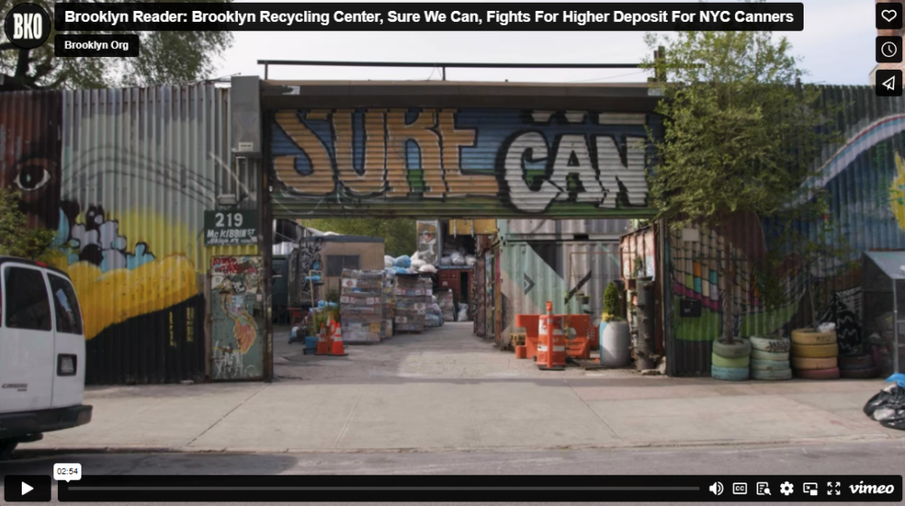 Entrance of Brooklyn recycling center "Sure We Can," showing stacks of recyclables and street art on the walls. Orange traffic cones and greenery are visible near the gate.