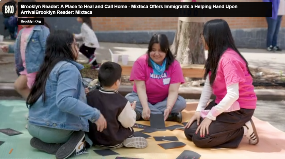 A group of people, including children, sit on a colorful blanket outdoors, engaging in an activity with black rectangular objects. Adults wear pink and blue Mixtca shirts, with a brick wall in the background.