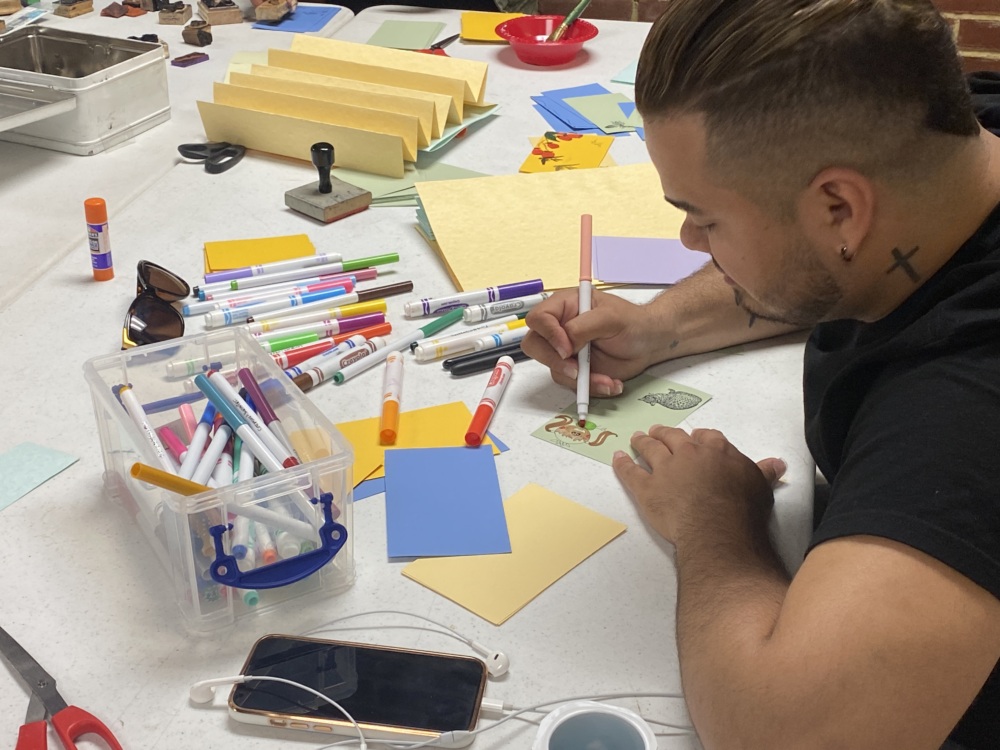 A person with a tattoo on their neck is drawing on a piece of paper at a table filled with markers, glue sticks, a smartphone, and other craft materials.