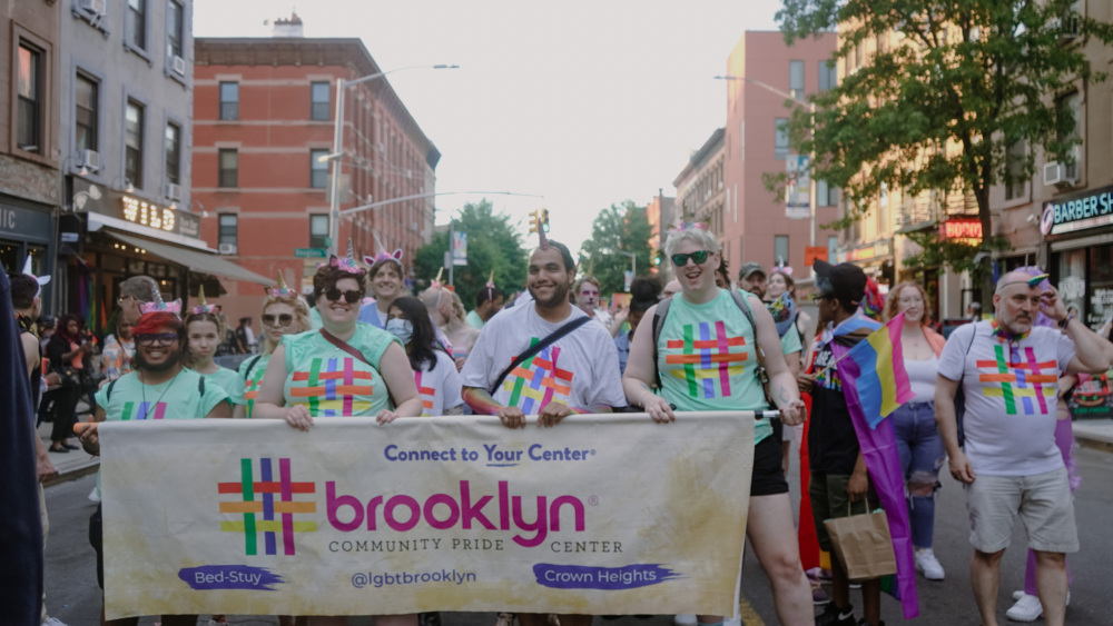 A group of people wearing matching shirts and holding a banner that reads "Brooklyn Community Pride Center" walk in a pride parade down a street lined with buildings and trees.