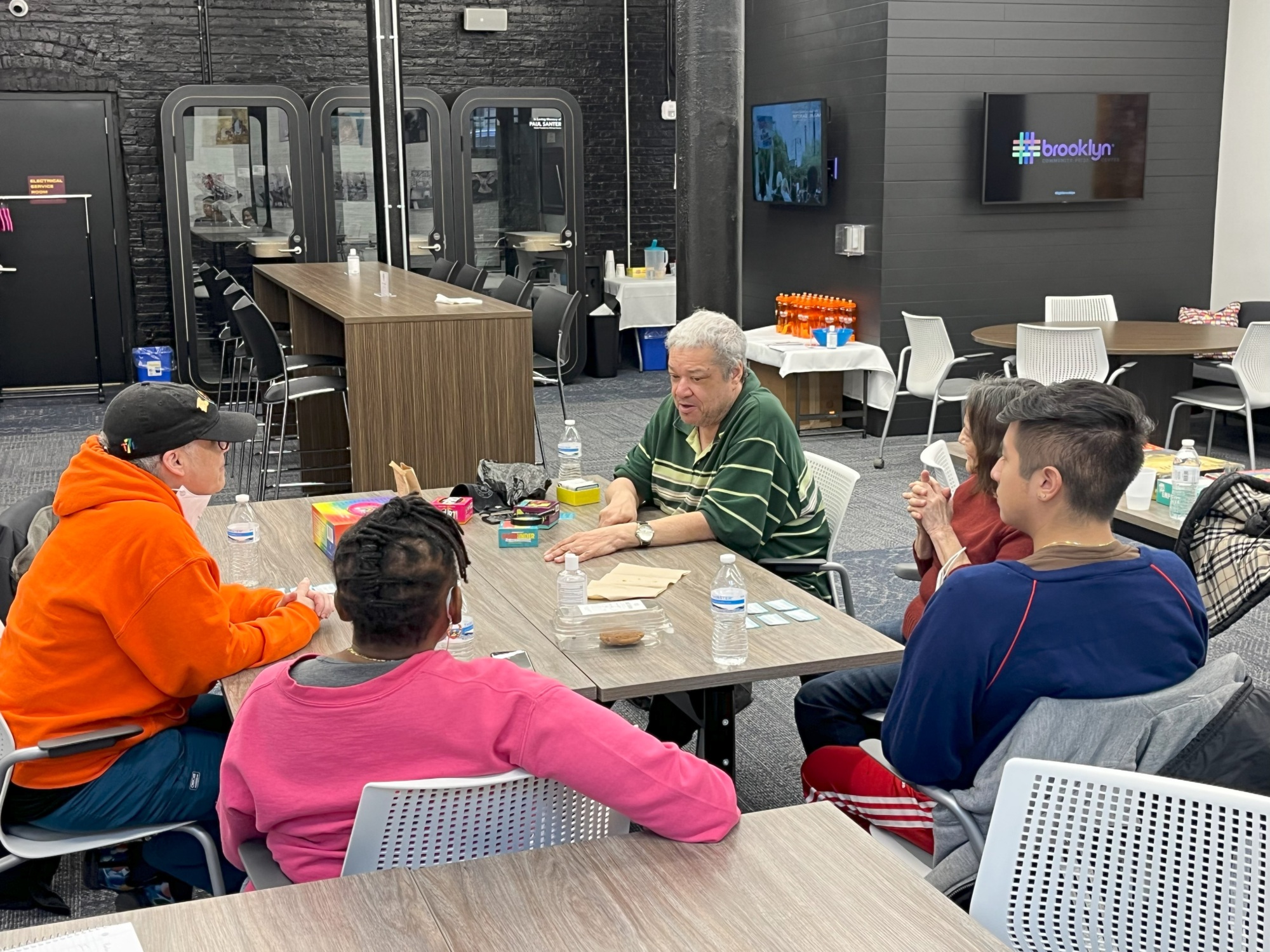 A group of people sit around a table in a modern indoor space, engaged in conversation. Bottled water and various items are on the table. A display on the wall reads "#brooklyn".